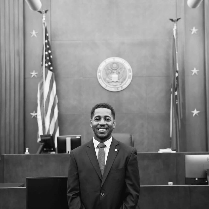 A smiling man in a suit stands in the foreground of a courtroom featuring an American flag and a government seal in the background.