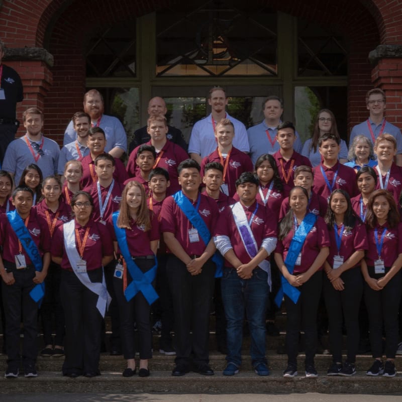 A large group of students and adults in maroon shirts pose for a photo in front of a brick archway.