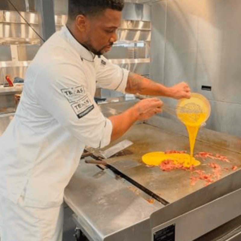 A chef in a white uniform pours yellow batter from a strainer onto a flat grill inside a professional stainless steel kitchen.