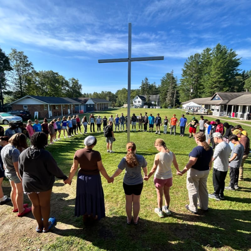 A group of people hold hands in a large circle around a tall metal cross in a grassy field, with small buildings and green trees under a blue sky in the background.
