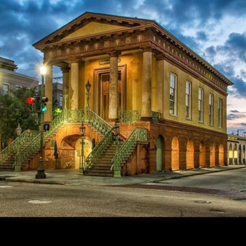 A grand, classical yellow building with ornate green staircases stands beside a paved street under a dramatic cloudy sky.