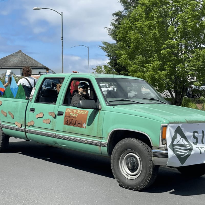A green pickup truck decorated with footprints and a sign is parked on a paved road with green trees and a building in the background.