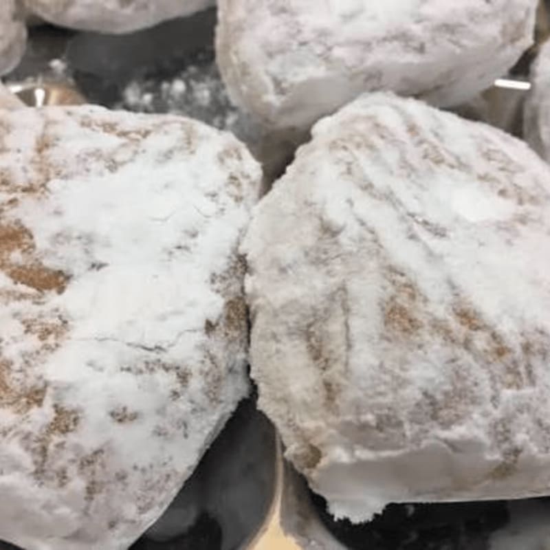 Powdered sugar-coated pastries sit in the foreground against a blurred background of more treats on a metallic tray.