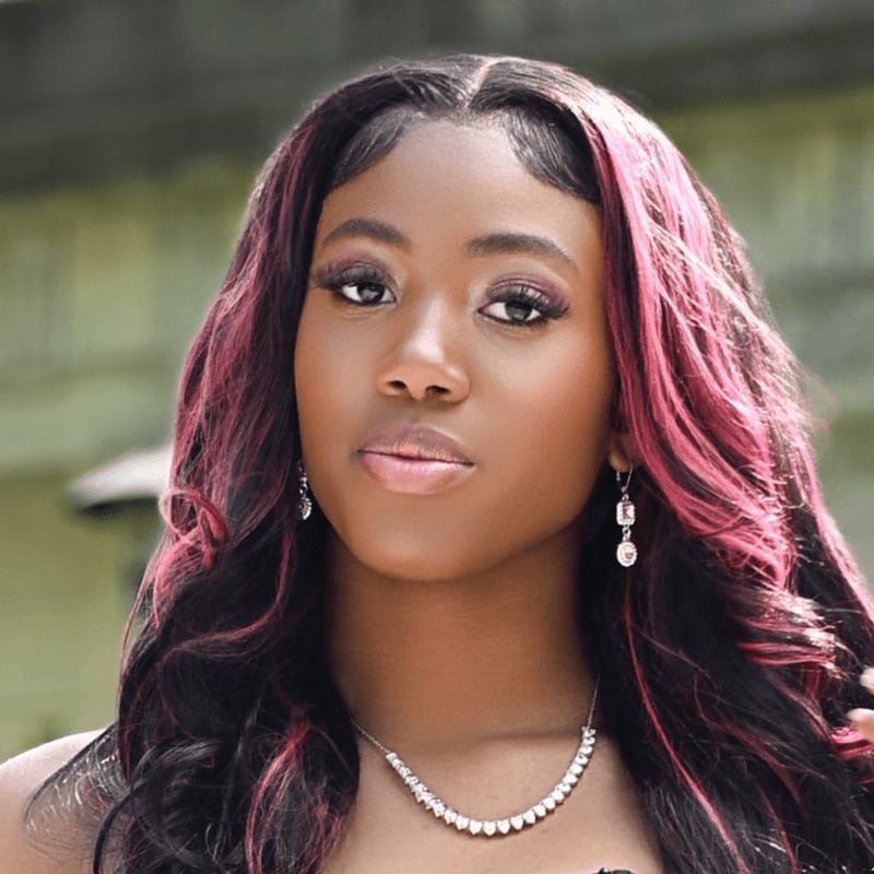 A woman with long black hair highlighted in pink and wearing a diamond necklace poses against a blurred, neutral-toned background.