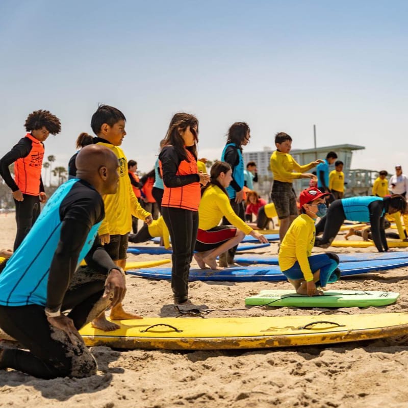 Children in colorful rash guards practice on surfboards on a sandy beach, with a pier and buildings visible in the background under a clear blue sky.