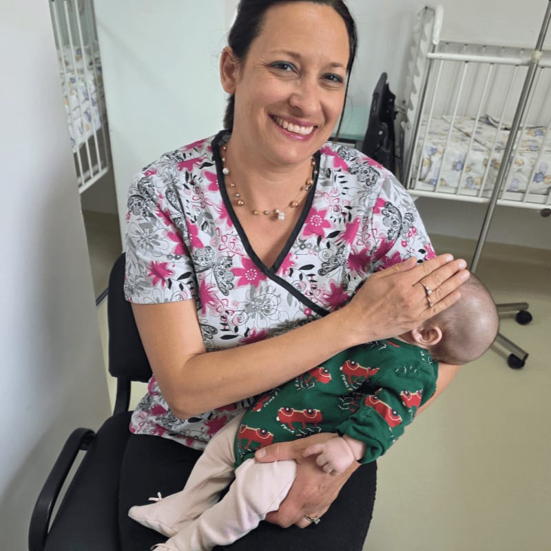 A smiling woman in a floral scrub top holds a baby in a green outfit while sitting in a room with a white medical crib in the background.