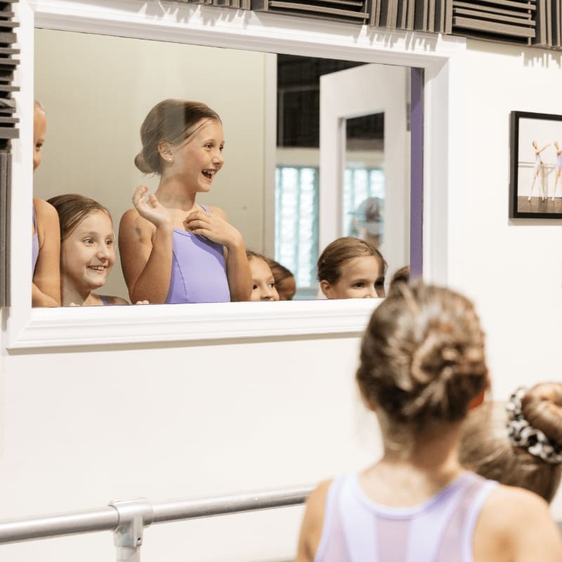 A young ballerina stands in the foreground facing a large mirror that reflects a group of smiling girls in a dance studio.