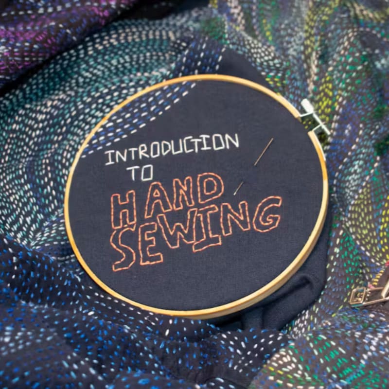 An embroidery hoop with the words "INTRODUCTION TO HAND SEWING" rests on a backdrop of colorful, textured fabric.