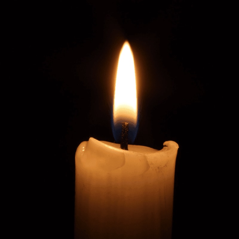 A lit white candle with a glowing yellow flame stands against a solid black background.