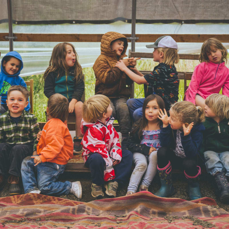 A group of young children in colorful jackets sit together on a wooden bench in the foreground, with a neutral-toned fabric backdrop behind them.