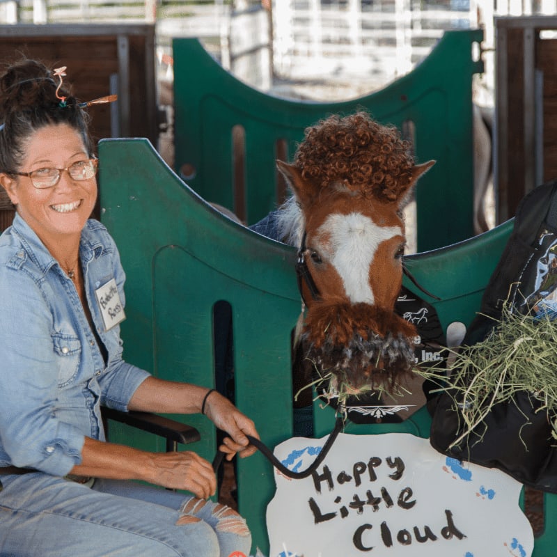 A smiling woman in a denim shirt holds a lead rope attached to a small horse with a curly mane, while a sign reading "Happy Little Cloud" sits in the foreground and a stable is visible in the background.