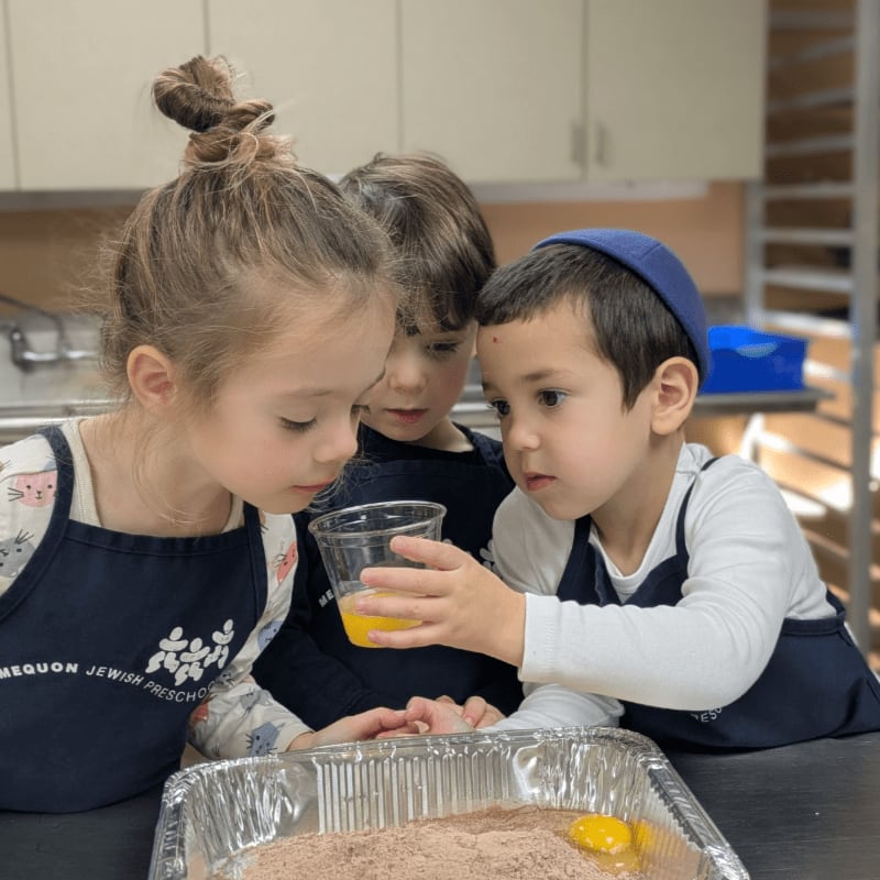 Three young children wearing aprons gather around a baking pan in a kitchen with white cabinets.