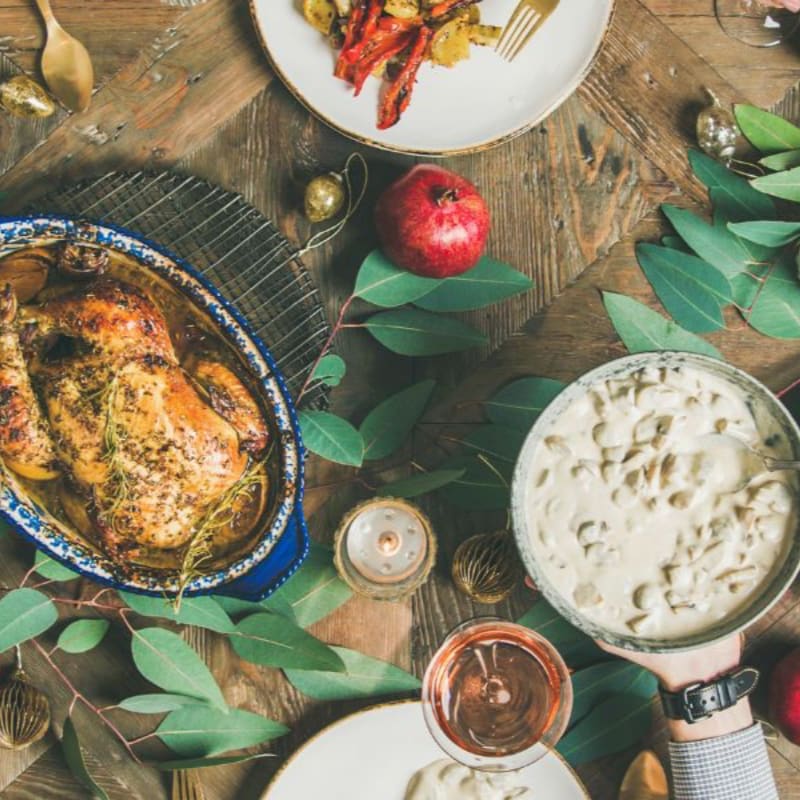 A roasted chicken and a bowl of creamy soup sit on a rustic wooden table adorned with green leaves, a pomegranate, and side dishes.