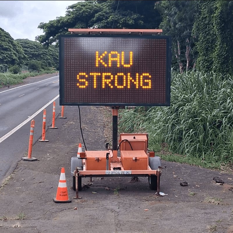 An electronic road sign reading "KAU STRONG" stands on a shoulder next to a road, with orange traffic cones in the foreground and lush green trees and grass in the background.