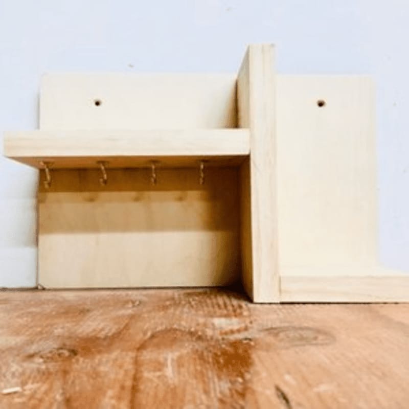 A light-colored wooden key holder with hooks is mounted on a white wall, with a wooden floor in the foreground.