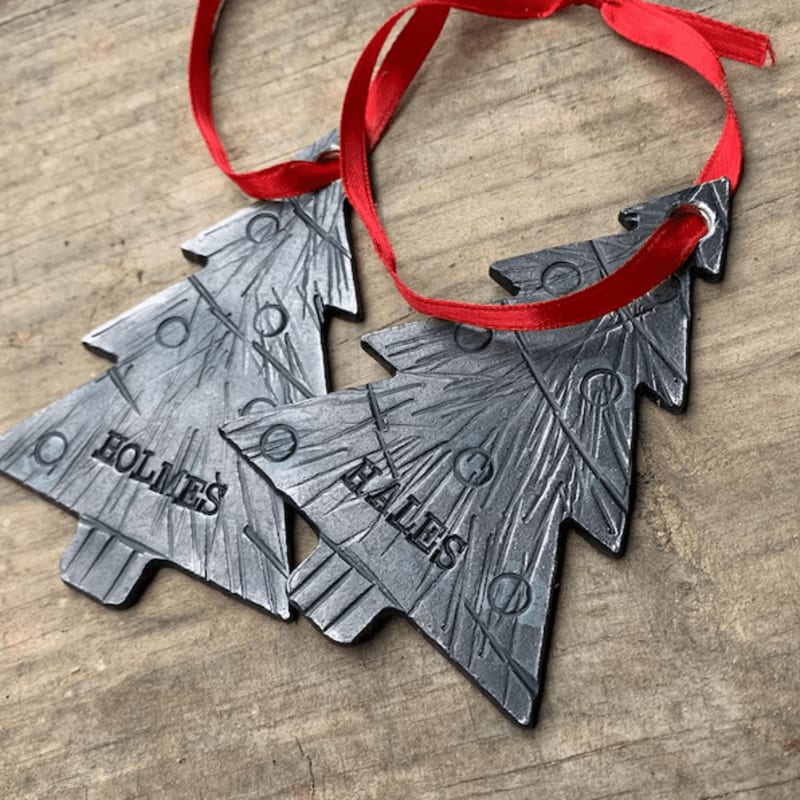 Two silver-colored tree-shaped ornaments with the names "HOLMES" and "HALES" and a red ribbon rest on a rustic wooden surface.