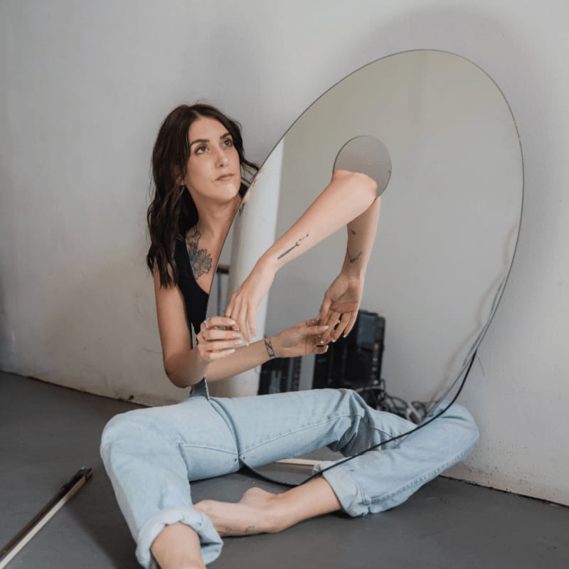 A woman in a black top and light blue jeans sits on a grey floor leaning against a white wall, posing with a large, irregularly shaped mirror.