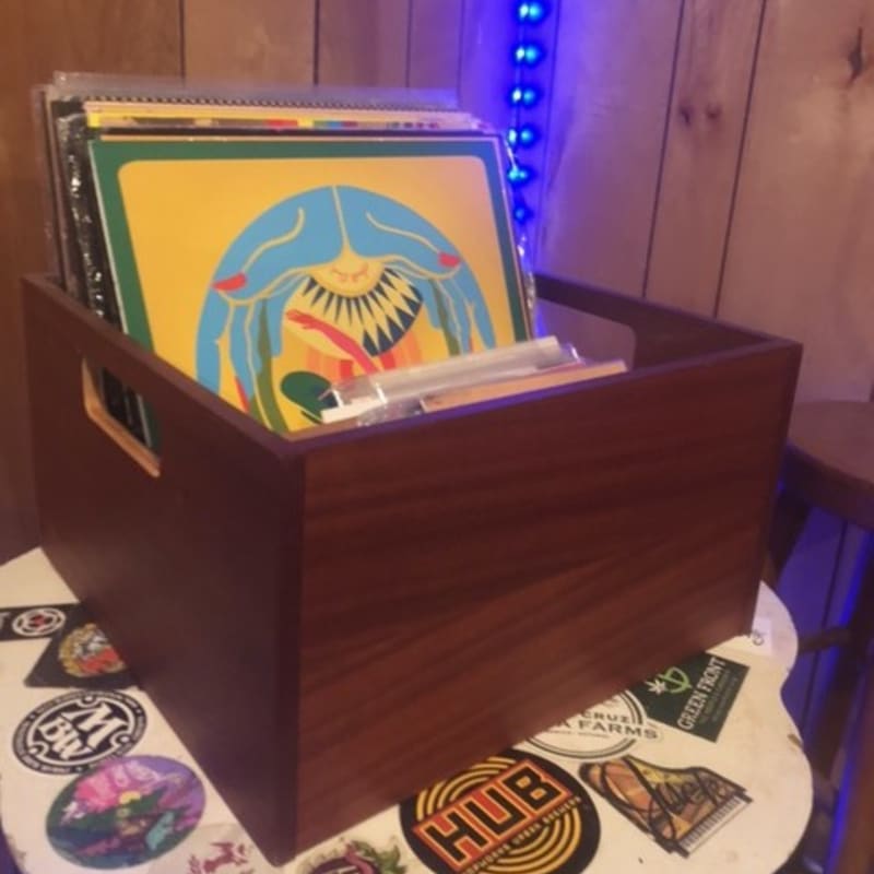 A wooden crate filled with vinyl records sits on a sticker-covered table against a wood-paneled wall with blue string lights.