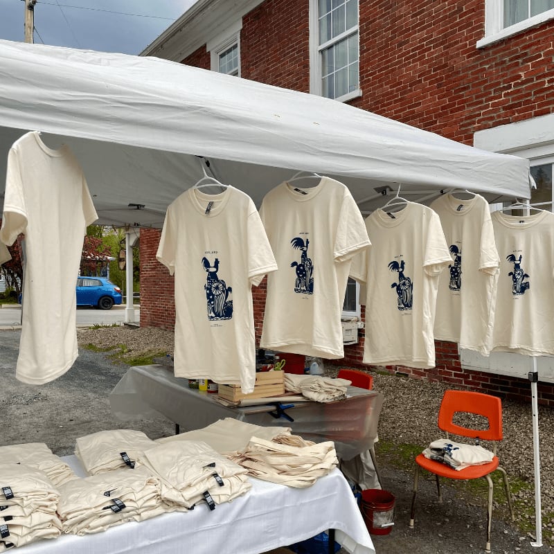 Cream-colored t-shirts with blue graphics hang under a white canopy in front of a red brick building.