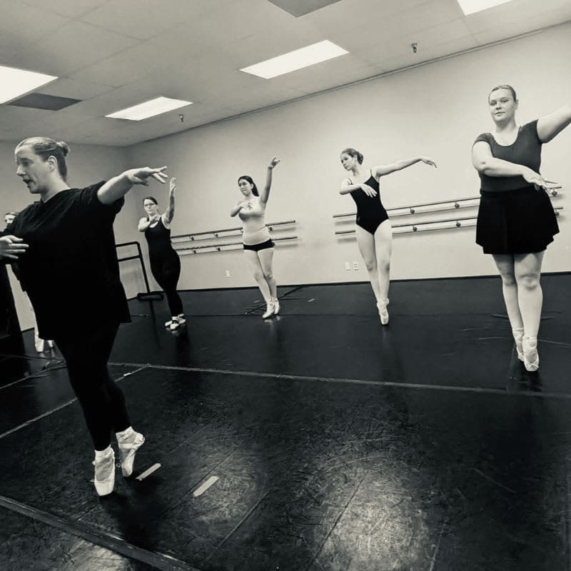 Several dancers in pointe shoes practice ballet in a studio with barre equipment in the background.