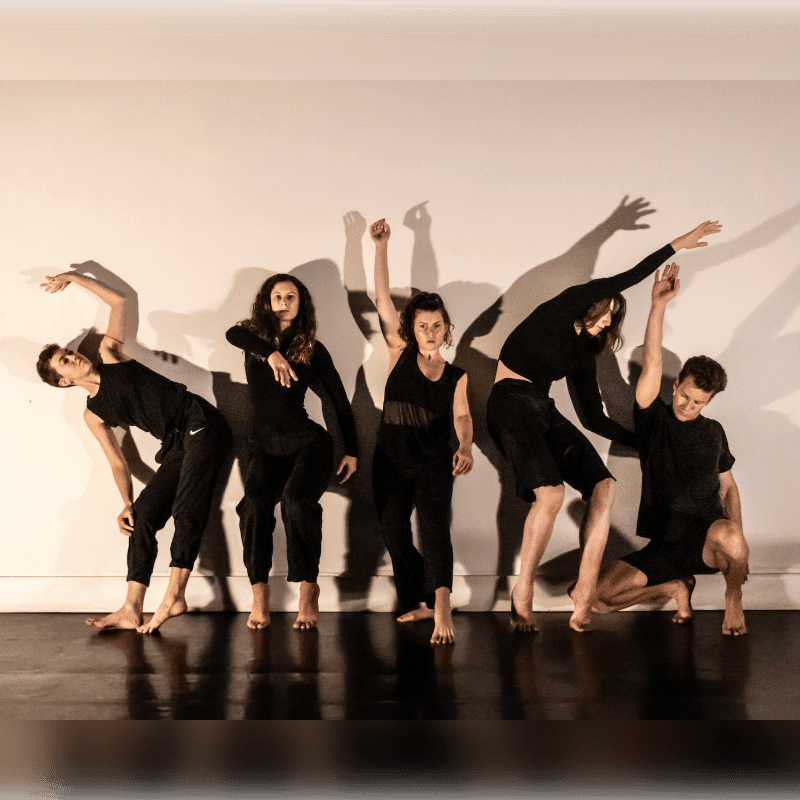 Five dancers in black attire perform expressive poses in the foreground against a plain white wall that captures their shadows in the background.