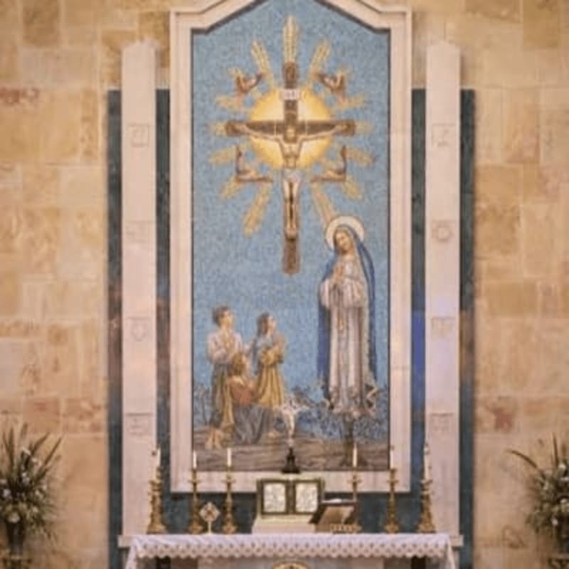 In the foreground, an altar is set with candles and books, while the background features a large mosaic depicting Jesus on the cross, Mary, and two children against a stone wall.