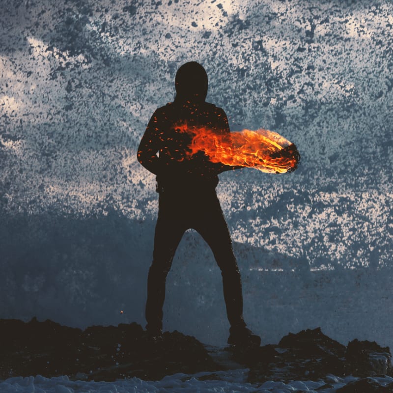 A silhouetted figure holds a blazing torch of fire while standing on a dark rocky shore against a backdrop of crashing white ocean spray.