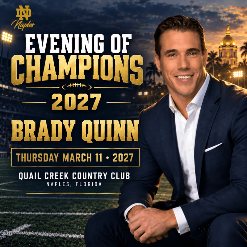 A smiling man in a navy blue suit sits in the foreground against a background of a football stadium and a golden dome under a twilight sky.
