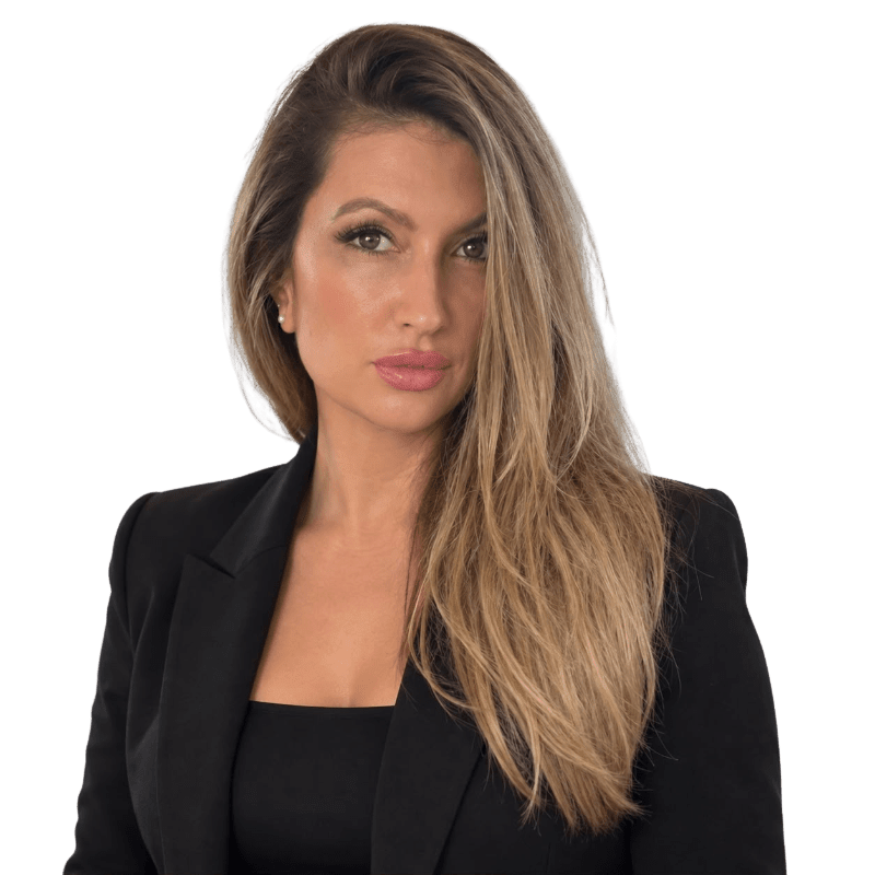 A woman with long blonde hair wearing a black blazer stands against a plain white background.