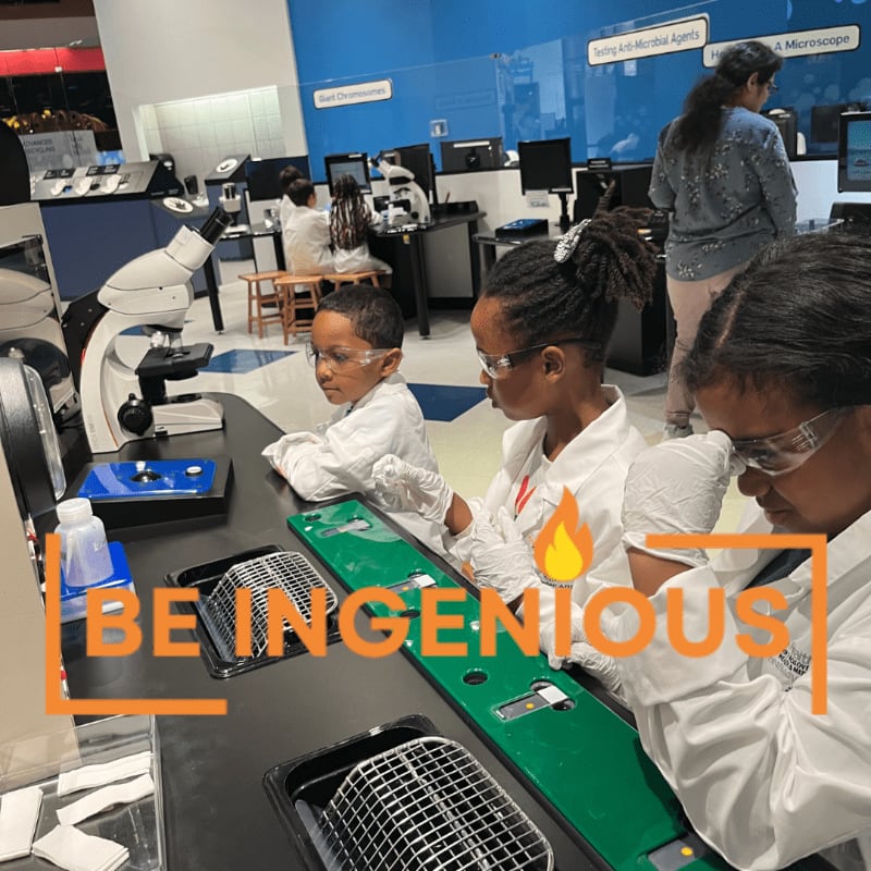 Children in white lab coats and safety goggles work at a science station in the foreground, while other students and a teacher are visible in a modern laboratory background.