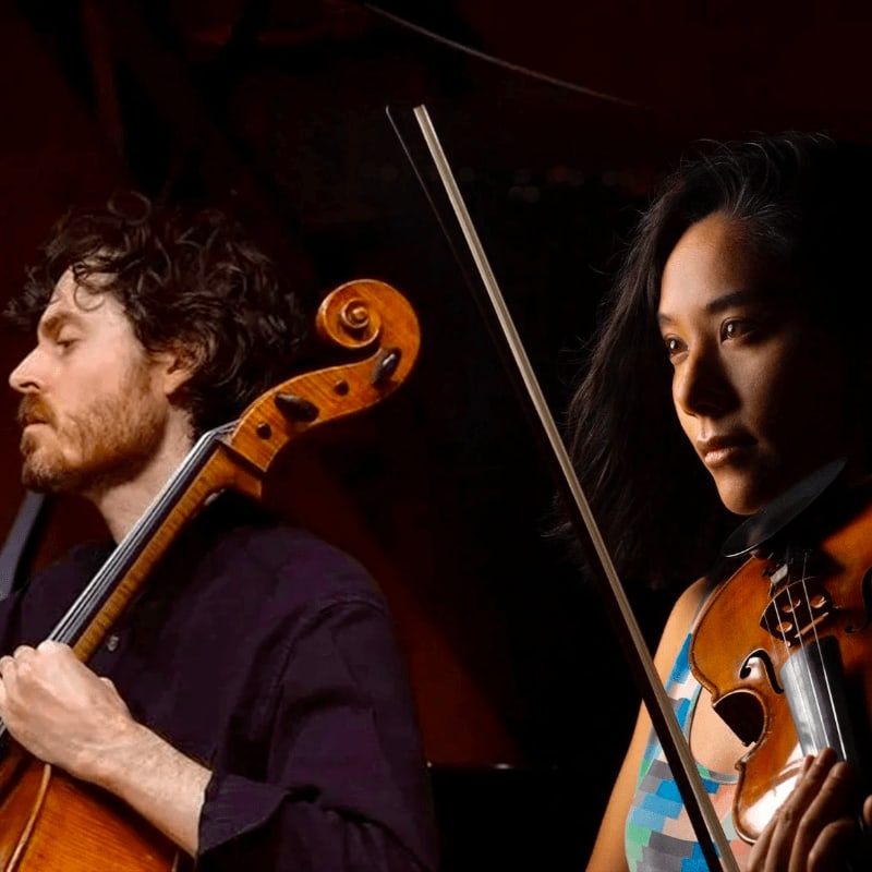 A man playing a cello and a woman holding a violin stand in the foreground against a dark, shadowed background.