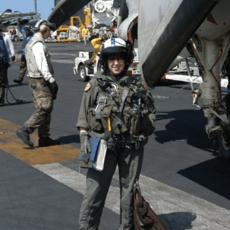 A pilot in full flight gear stands in the foreground on an aircraft carrier deck with a jet and crew members in the background.