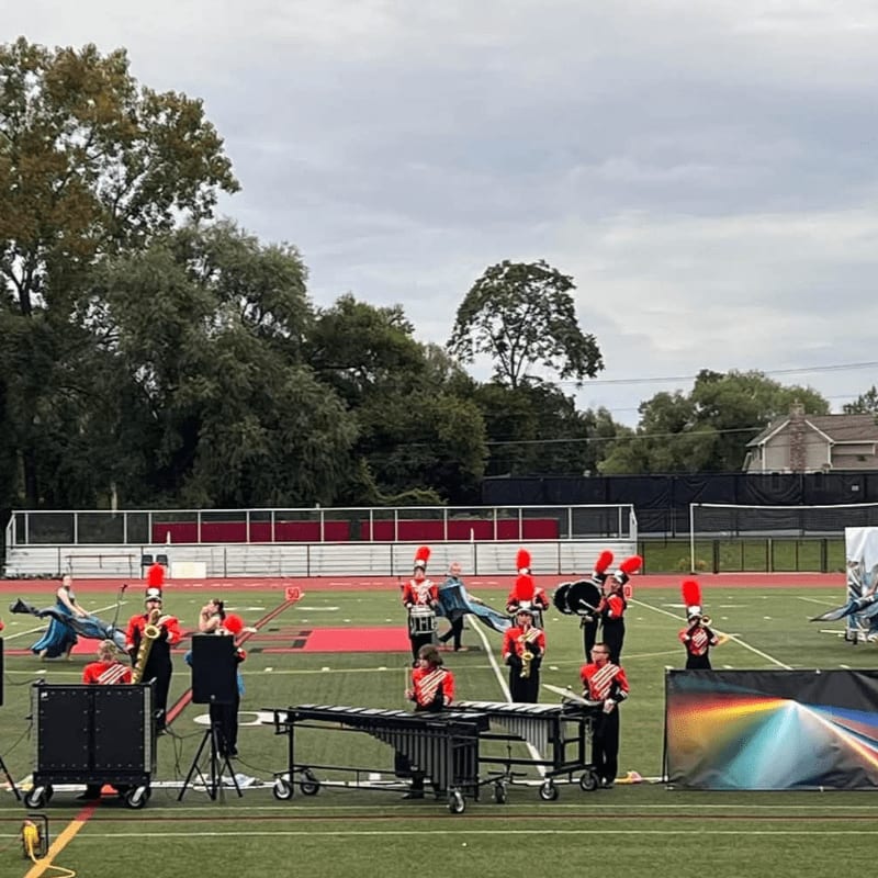 A marching band in red and black uniforms performs on a green football field against a backdrop of lush green trees and a cloudy sky.