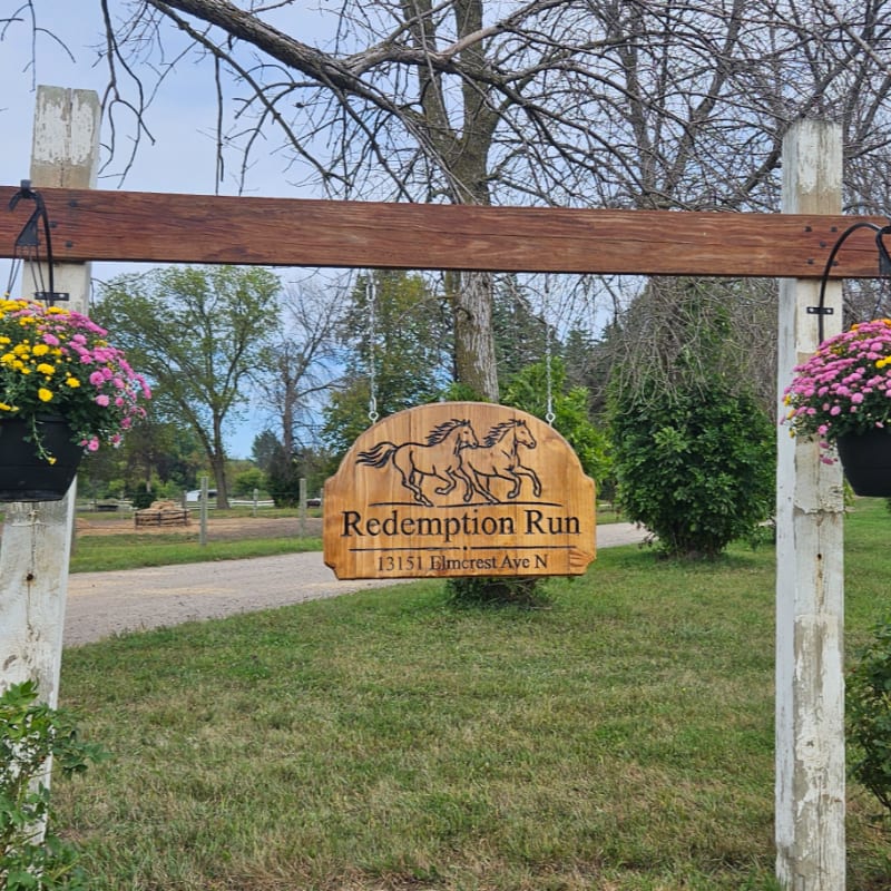 A wooden "Redemption Run" sign hangs from a rustic beam between two white posts and flower baskets, with a grassy path and leafy trees in the background.