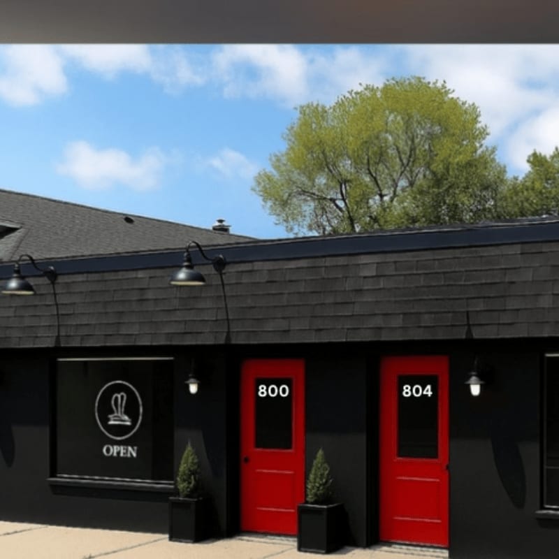 A black building with two red doors and an "OPEN" sign stands under a blue sky with a large green tree in the background.