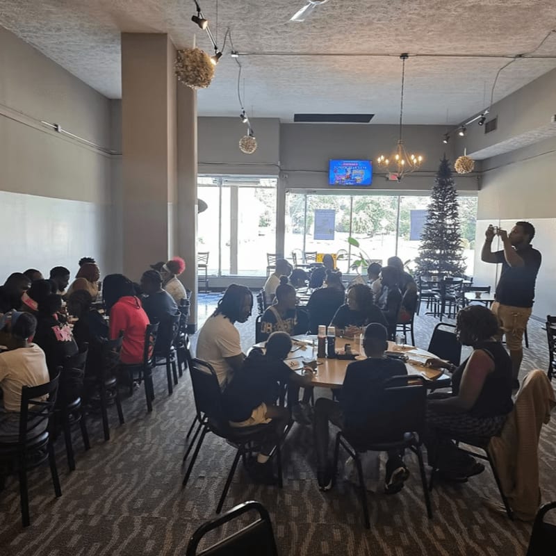 A group of people sit around tables in a modern room with a television and a Christmas tree in the background.