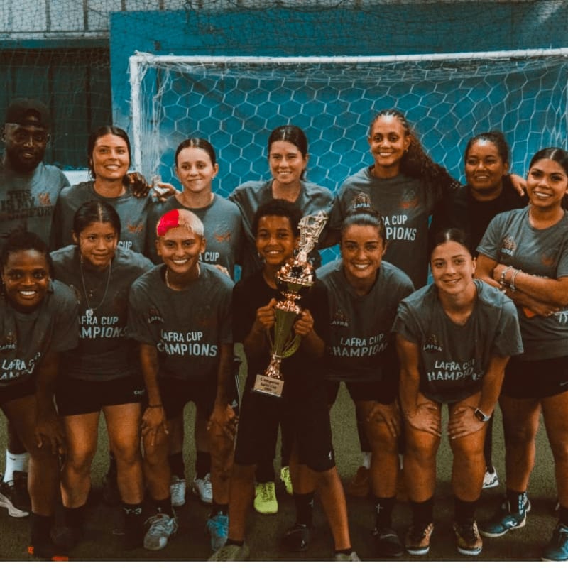 A group of female soccer players in grey shirts pose with a trophy in the foreground against a blue wall and goal net in the background.