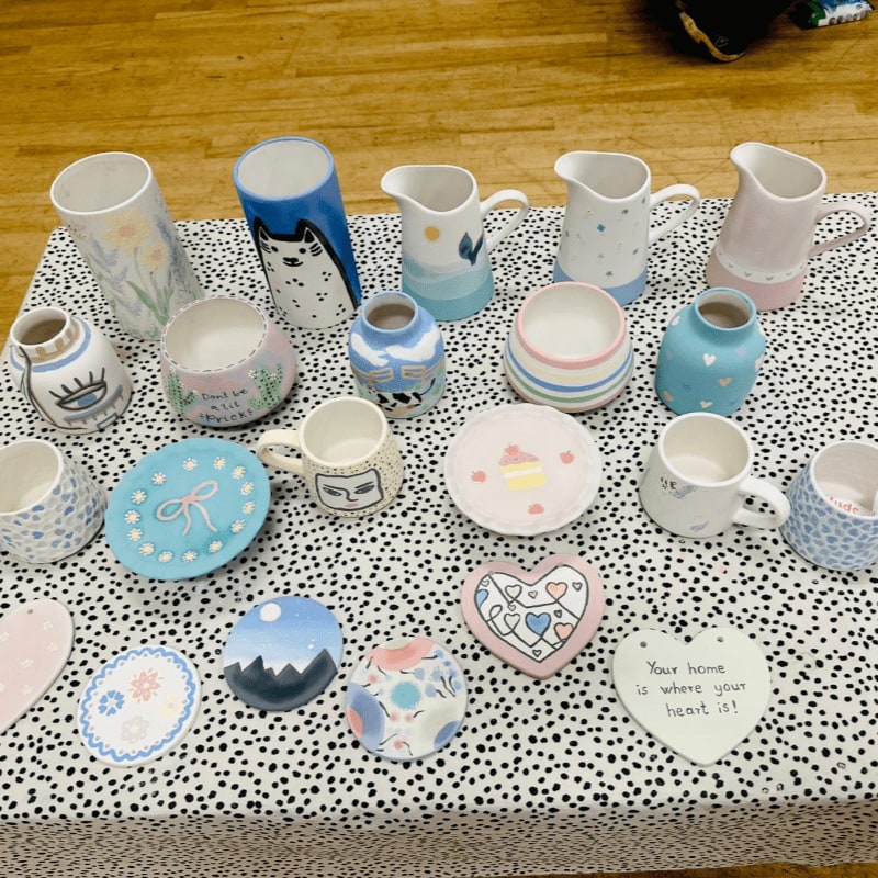 A variety of hand-painted ceramic mugs, pitchers, and heart-shaped dishes are arranged on a black-and-white dotted tablecloth over a wooden floor.