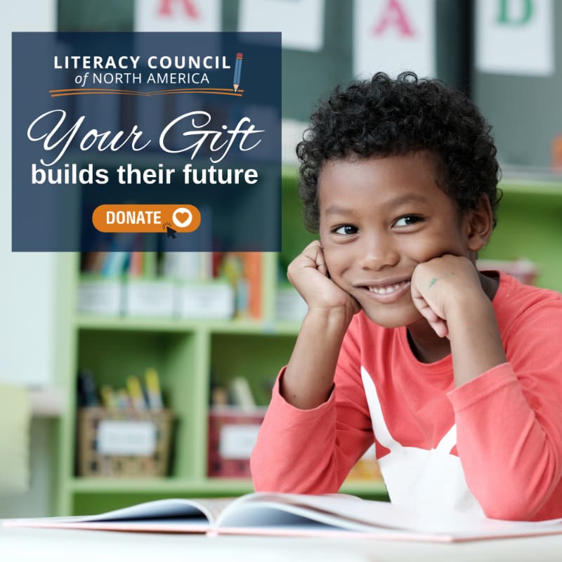 A smiling young boy in a red shirt rests his chin on his hands over an open book, with a blurred classroom background and a donation overlay in the upper left.