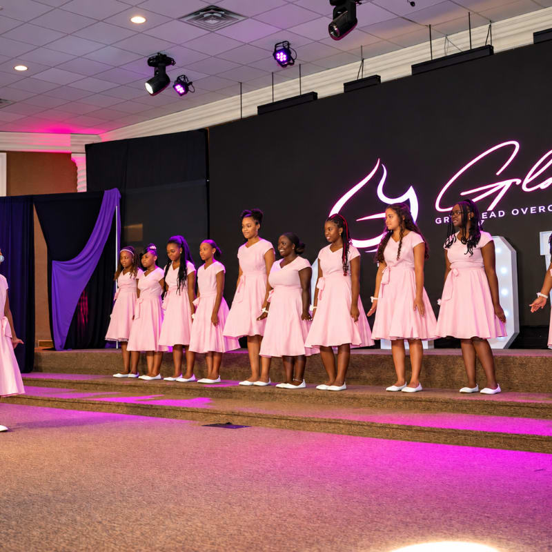A line of young girls in light pink dresses stands on a stage in front of a large black backdrop.