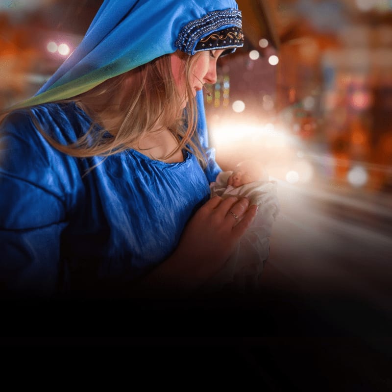 A woman in a blue headscarf and dress cradles a glowing infant against a blurred, warm-toned background of bokeh lights.