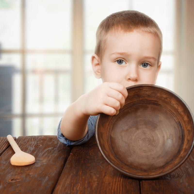 A young boy holds a large wooden bowl in front of his face while sitting at a rustic wooden table against a blurred indoor background.