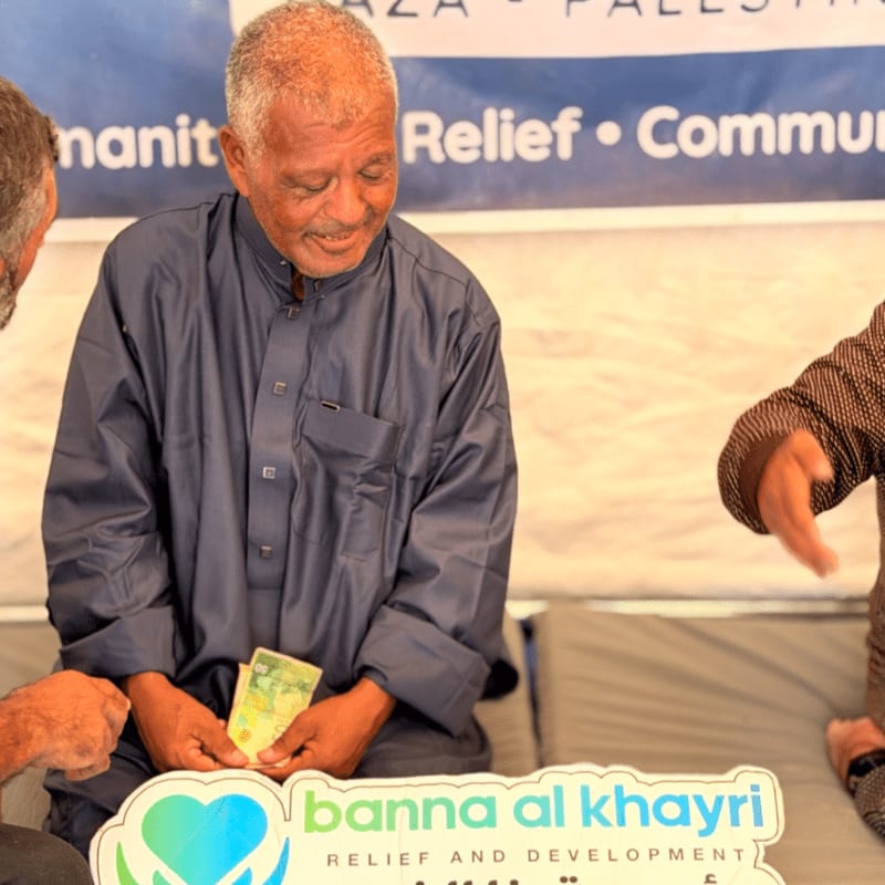 An elderly man in a dark blue robe holds money in the foreground, set against a background featuring a blue and white relief organization banner.