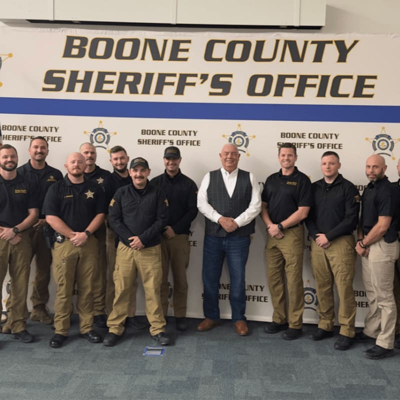 A group of law enforcement officers and a man in a vest stand in the foreground against a background featuring a large "Boone County Sheriff's Office" banner.