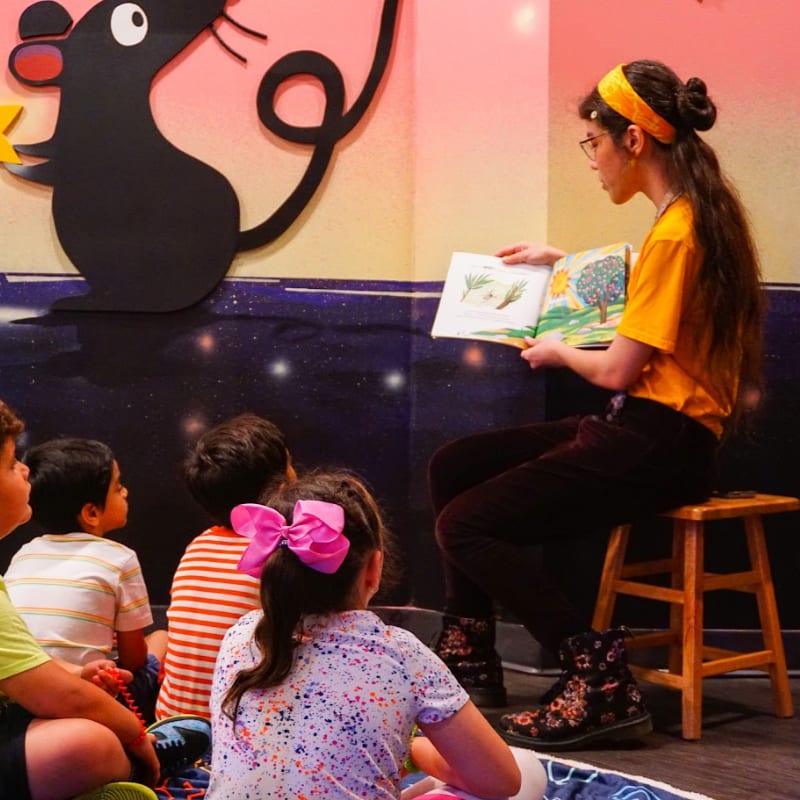 A woman in a yellow shirt reads a book to a group of children sitting on the floor in front of a wall featuring a large black mouse illustration.