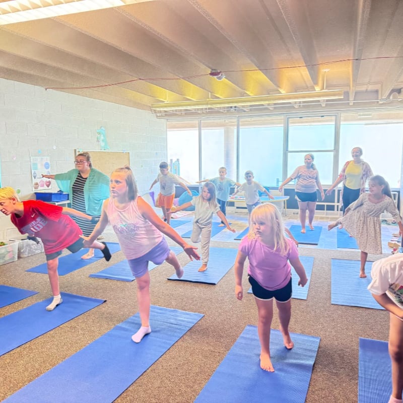 Children practice yoga on blue mats in a bright room with a teacher and large windows in the background.