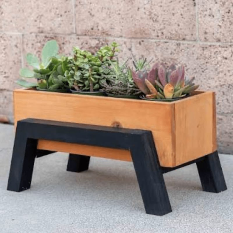 A wooden planter box filled with succulents sits on a black stand against a textured stone wall.