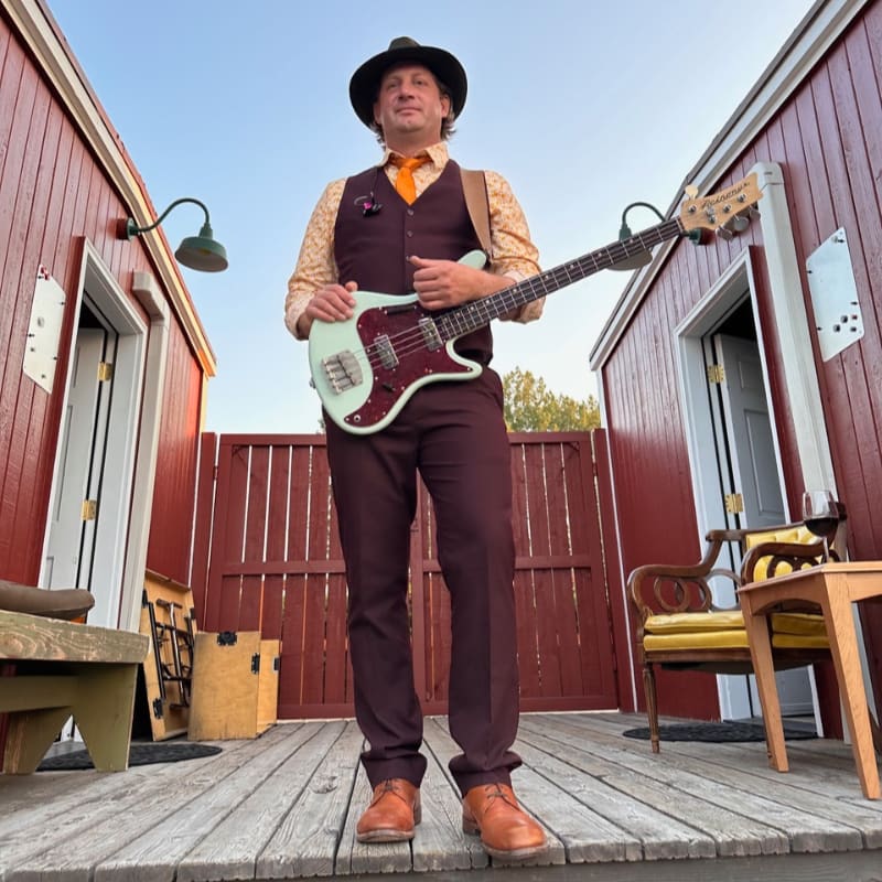 A man in a vintage vest, hat, and tie holds a light-colored bass guitar while standing on a wooden deck between two red buildings.
