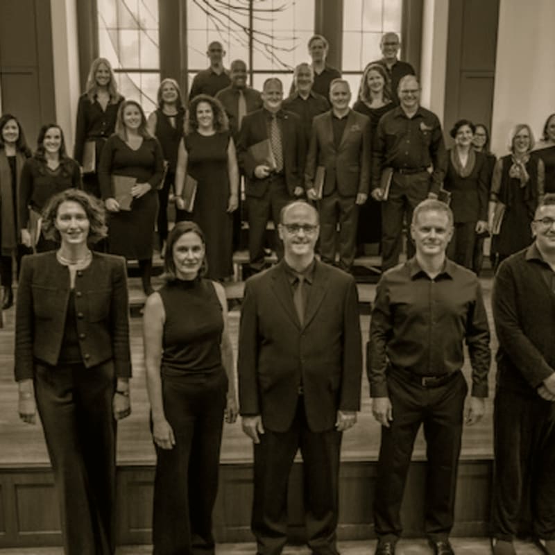 A group of people in formal black attire pose for a tiered group photo inside a room with large windows in the background.
