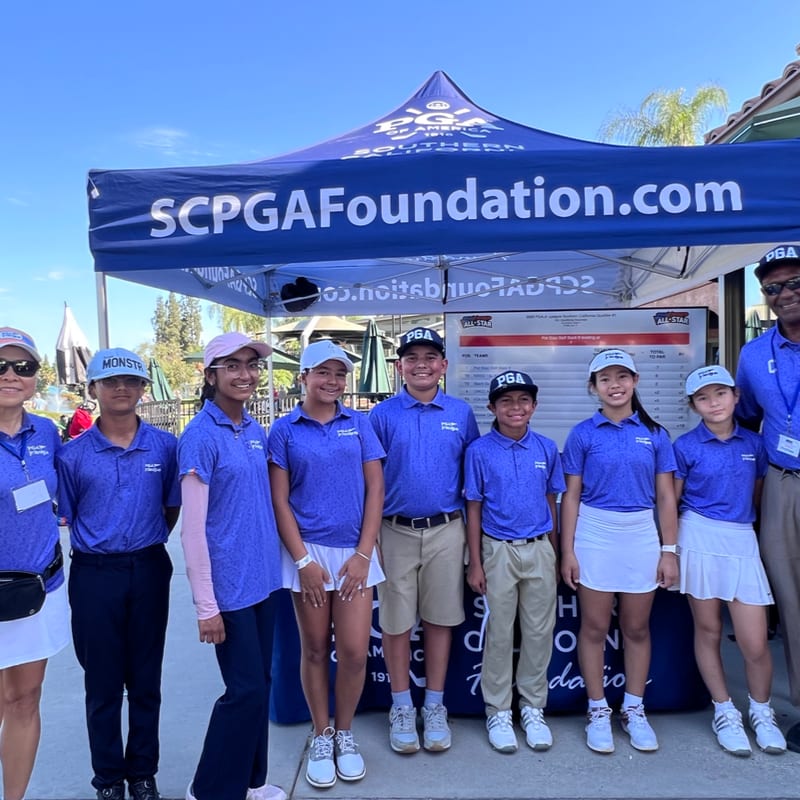 A group of young golfers in blue shirts pose together in the foreground in front of a blue SCPGA Foundation tent and a clear blue sky.
